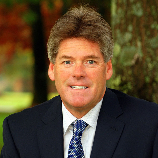 Portrait of a middle-aged man in a navy suit and blue tie, outdoors with trees in the background.